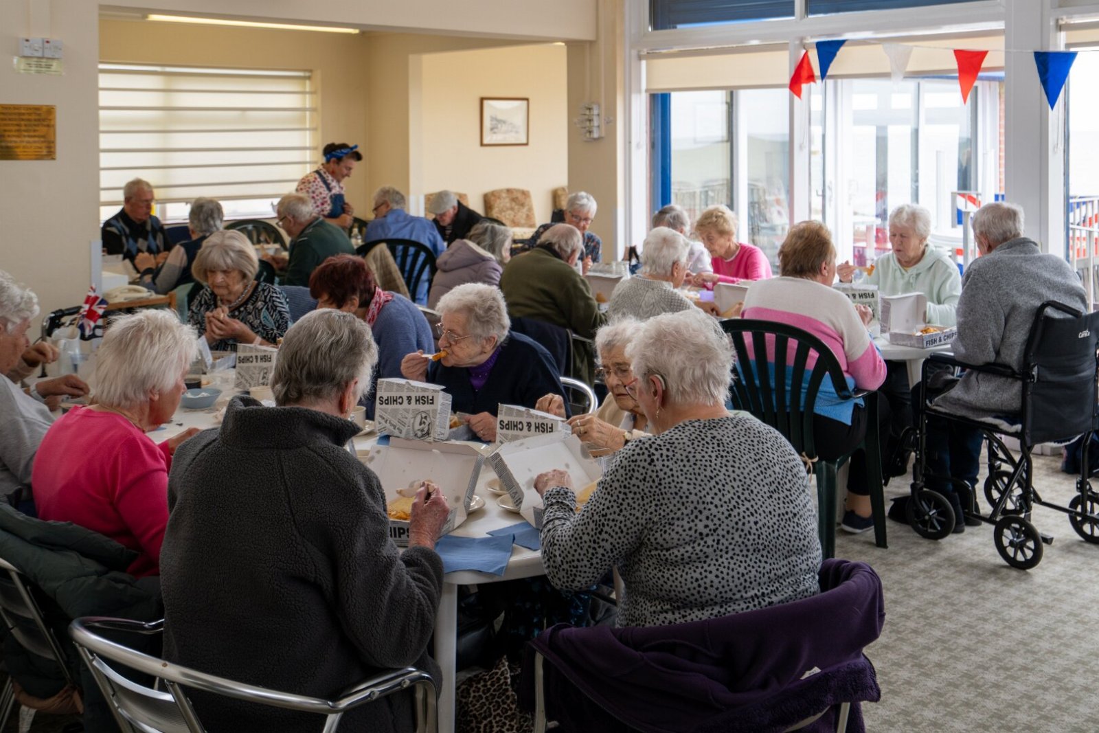 A group of residents sitting at tables inside a bright seaside venue, enjoying fish and chips from traditional boxes during a community outing.