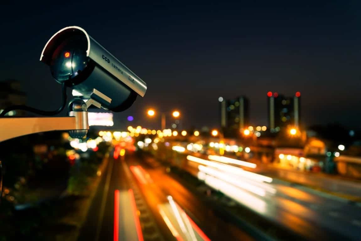 CCTV camera mounted on a lamp-post beside a well-lit residential street in a UK town at dusk.