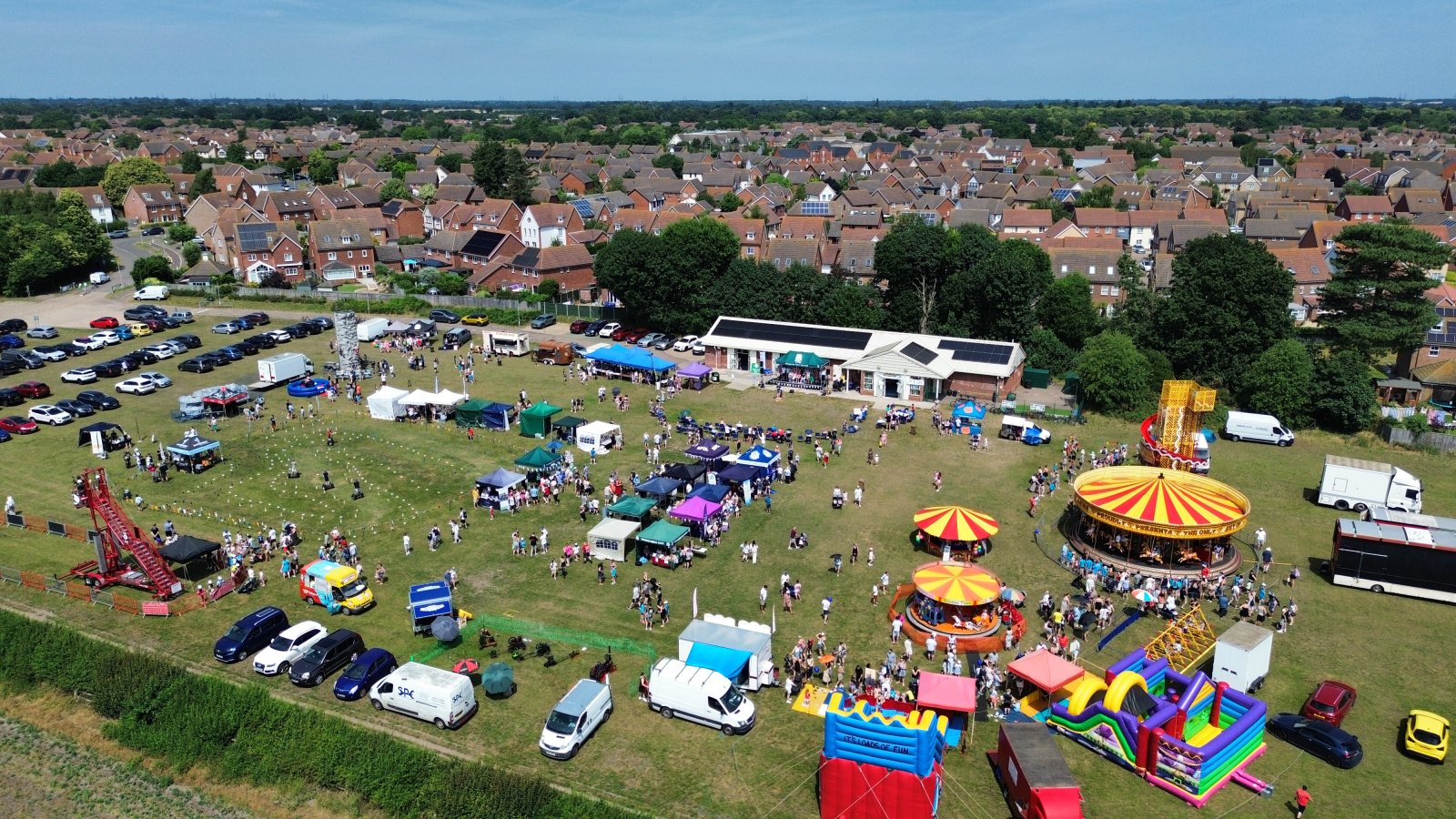 A wide, sunny shot of the Kesgrave Fun Day at Millennium Sports Field, showing families enjoying fairground rides like a golden carousel and a tall spiral slide under a clear blue sky.